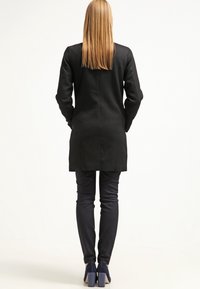 Pitkä musta takki, jossa suora malli ja sileä tekstuuri. Yhdistetty tummiin housuihin ja sinisiin korkokenkiin.
