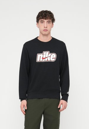 Czarna bluza z nadrukiem logo Nike w kolorach białym i czerwonym, z okrągłym dekoltem, długimi rękawami i ściągaczami przy mankietach, zestawiona z oliwkowozielonymi spodniami typu cargo.