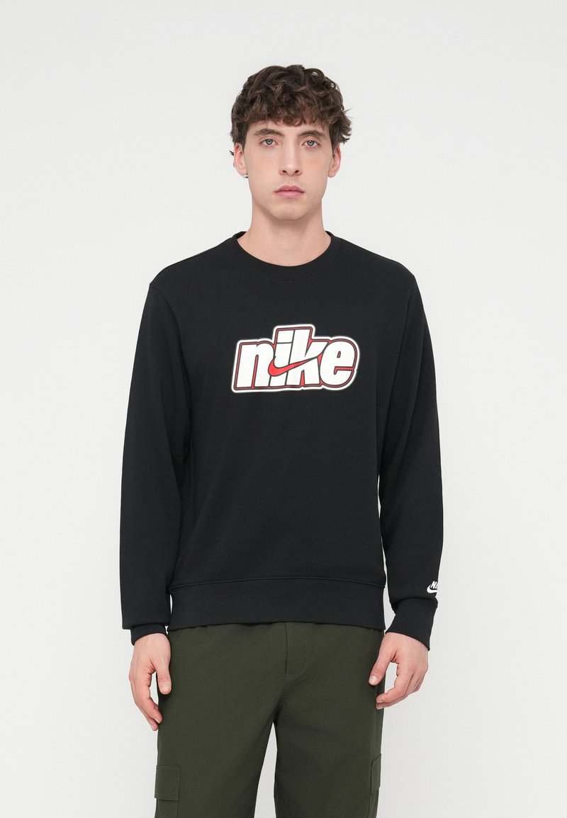 Čierna mikina s bielym a červeným logom Nike, s okrúhlym výstrihom, dlhými rukávmi a rebrovanými manžetami, v kombinácii s olivovo zelenými vojenskými nohavicami.