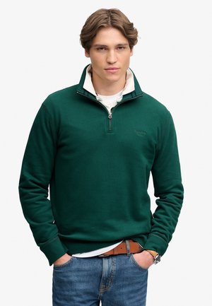 Junger Mann mit hellbraunen Haaren, der einen dunkelgrünen Sweatshirt mit Viertel-Reißverschluss, ein weißes Hemd darunter, blaue Jeans und einen braunen Gürtel trägt.