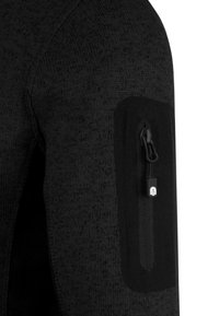 Schwarze gestrickte Jackenärmeln mit einem strukturierten Muster. Verfügt über ein schwarzes Stoffpatch und eine mit einem Ziehband gesicherte Reißverschlusstasche, die fest angenäht ist.