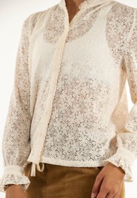 Blusa in pizzo di un bianco sporco con colletto sagomato, motivo floreale intricato, chiusura con bottoni e polsini arricciati. Tessuto leggero e semi-trasparente.