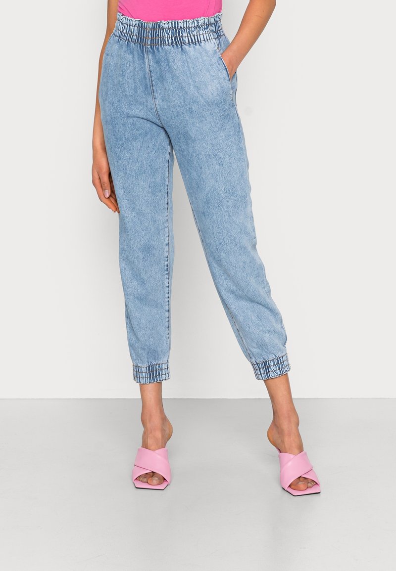 Femme portant un pantalon de jogging en denim bleu clair à taille élastique et avec des poignets, ainsi que des sandales à talons roses, se tenant devant un fond blanc.