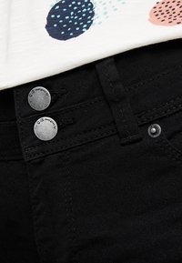 Pantalones vaqueros de mezclilla negra con botones metálicos, que presentan un primer plano de la cintura y costuras intrincadas. Tejido texturizado y detalles mínimos.