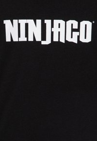 Schwarzes Baumwoll-T-Shirt mit großen weißen Blockbuchstaben, die "NINJAGO" über die Brust schreiben. Einfaches Design ohne zusätzliche Grafiken.