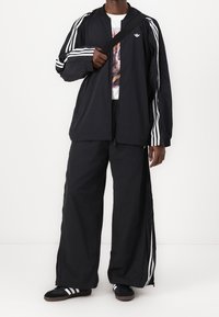 Svart träningsdräkt med zip-up jacka och vida byxor, båda med vita ränder längs armarna och sidorna, tillverkad av lättviktig tyg.