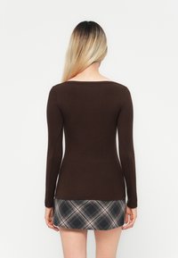 Pikkade blondide juustega naine, seljas vormiv pruun pikka varrukatopp ja must-valge ruuduline mini seelik, seisab valge tausta ees.
