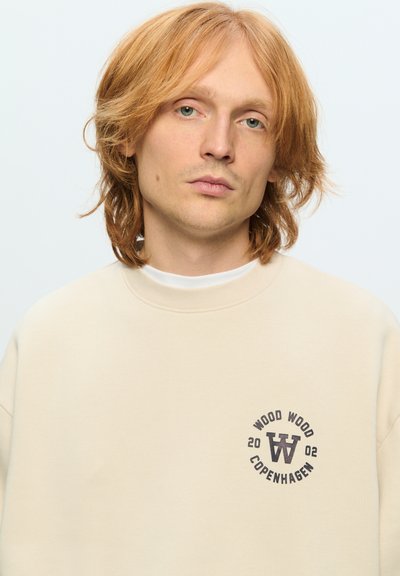 Sweatshirt beige avec un logo rond présentant "WOOD WOOD COPENHAGEN" et un "W" stylisé. Texture douce, design col rond, style minimaliste.