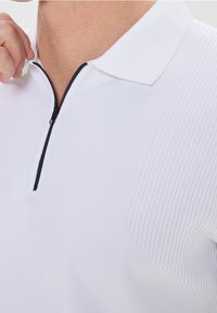 Osoba upravující černý zip na bílém pleteném polokošili s krátkým rukávem a límečkem, která má texturovaný detail na ramenech.