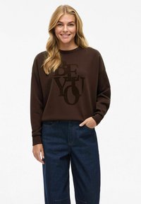Sweatshirt marron avec un graphique "SOIS TOI" en un matériau doux et pelucheux ; associé à un jean large bleu foncé ; coupe décontractée avec des manches relaxées.