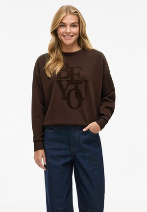Sweatshirt marron avec un graphique "SOIS TOI" en un matériau doux et pelucheux ; associé à un jean large bleu foncé ; coupe décontractée avec des manches relaxées.