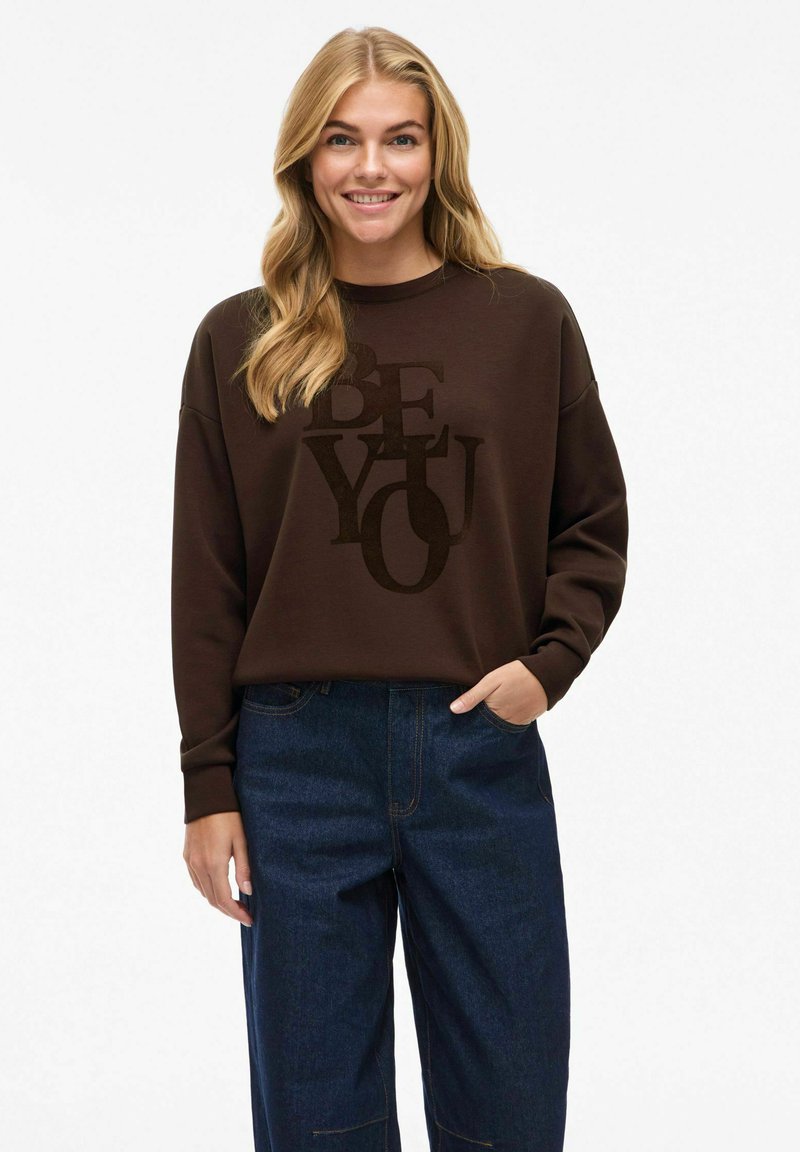 Sweatshirt marron avec un graphique "SOIS TOI" en un matériau doux et pelucheux ; associé à un jean large bleu foncé ; coupe décontractée avec des manches relaxées.