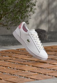 Baskets blanches Lacoste avec un accent rose au talon, flottant au-dessus de carreaux en bois en extérieur, avec des plantes floues et un mur en arrière-plan.