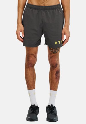 Kurze graue Shorts mit Gummibund und gelbem "ATW"-Aufdruck auf der Vorderseite, kombiniert mit dunklen Schuhen und weißen Socken mit goldener Schrift.