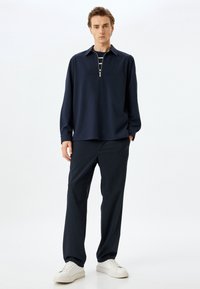 Dunkelblaues Langarm-Polo mit einem Viertelreißverschluss, darunter ein gestreiftes Hemd, kombiniert mit dunklen Hosen und weißen Sneakers. Glatte Textur.