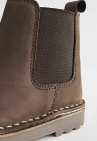 Stivaletto in suede marrone con pannelli laterali elastici, cuciture visibili e suola testurizzata. La parte superiore ha una finitura liscia e bordi rinforzati.