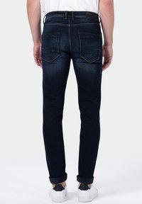 Un homme portant un jean bleu foncé retroussé et des baskets blanches, se tenant de dos contre un fond blanc.