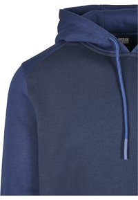 Felpa con cappuccio in tessuto blu scuro, caratterizzata da una finitura testurizzata e un pannello spalla a contrasto. Include cordini regolabili sul cappuccio.