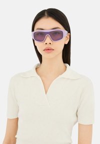 Frau mit glatten, dunklen Haaren, die übergroße violett getönte Sonnenbrillen und ein weißes kurzärmeliges Hemd mit Kragen vor einem schlichten Hintergrund trägt.