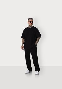 Schwarzes, übergroßes V-Ausschnitt-T-Shirt und passende Hose. Der Stoff scheint weich zu sein; das Model trägt weiße Turnschuhe und Sonnenbrille. Tätowierungen sind an den Armen sichtbar.