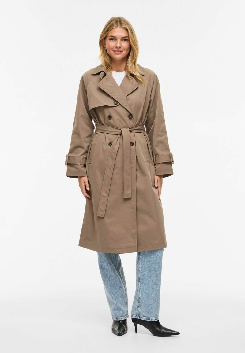 Trench beige à double boutonnage, taille ceinturée et grands revers. Associé à un jean délavé clair et des escarpins noirs à bouts pointus.