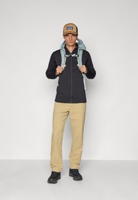 Black zip-up jacket with textured fabric, light blue backpack, beige pants, and black shoes; brown cap with logo detail.