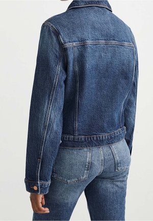 Osoba v tmavě modré džínové bundě a světlejších modrých džínových kalhotách, zobrazena ze zad na jednobarevném světle pozadí.