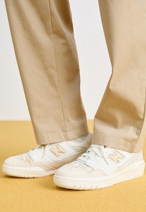 Pantalon beige porté avec des baskets blanches et beiges sur un sol marron clair.