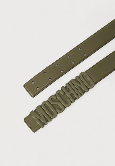 Ζώνη ελιάς με πολλαπλές τρύπες και μεγάλα γράμματα της επωνυμίας MOSCHINO ως αγκράφα σε απλό φόντο.