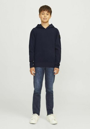 Felpa blu navy con tasca frontale e polsini a coste, abbinata a jeans scuri e sneakers bianche. Design semplice e pulito.