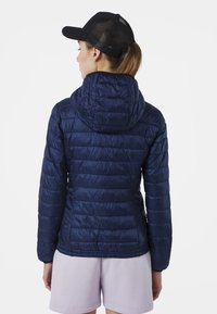 Giacca a vento blu navy con cappuccio, caratterizzata da cuciture orizzontali e una texture liscia e lucida. Indossata con pantaloni corti viola chiaro e un cappello nero.