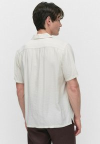 Chemise à manches courtes en tissu beige clair, texturé avec un léger froissement, dotée d'un col et d'une coupe décontractée. La vue de dos montre des plis et des fentes.