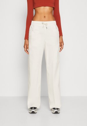 Pantaloni largi de culoare bej deschis, cu un cusătură ajustabilă la talie, având buzunare laterale și un logo subtil Nike pe piciorul stâng. Textură de țesătură netedă.