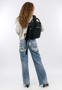 Svart ryggsäck med slät textur, flera blixtlåsförsedda fickor och justerbara remmar, bärs på ryggen av en modell i en stickad tröja och jeans.