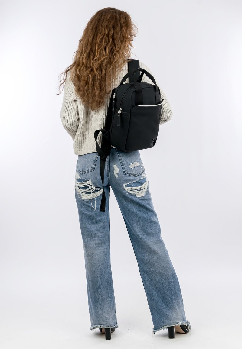 Svart ryggsäck med slät textur, flera blixtlåsförsedda fickor och justerbara remmar, bärs på ryggen av en modell i en stickad tröja och jeans.