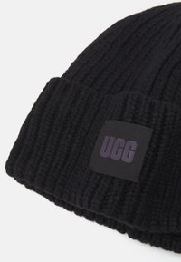 Beanie nero lavorato a maglia con texture a coste, caratterizzato da un'etichetta nera quadrata con la scritta "UGG" in lettering viola. Materiale morbido, con risvolto.