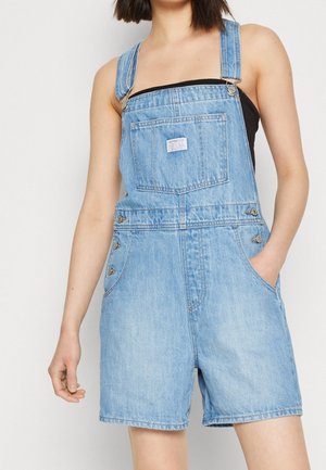 Jeune femme portant une salopette-short en jean bleu clair avec poche avant et un haut noir sans manches, main dans la poche, debout devant un fond blanc.