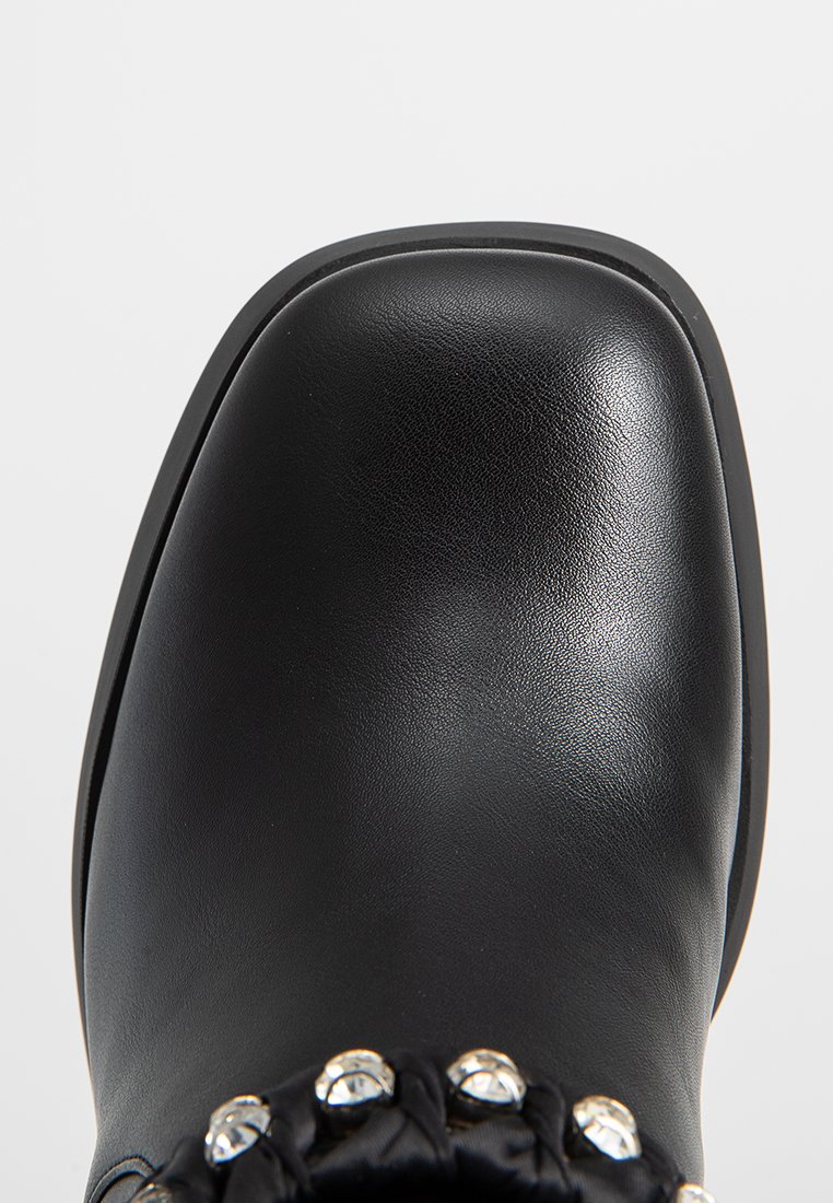 Bota de tobillo de cuero negro con punta redondeada, que presenta un acento de tela anudada y tachuelas metálicas a lo largo del cuello. Acabado mate, con un leve tacón.