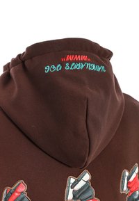 Sudadera marrón con texto bordado colorido en la capucha y parches de piernas en estilo de dibujos animados que llevan zapatillas en la parte posterior.