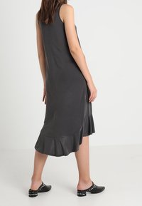 Szara sukienka bez rękawów z falbaną u dołu, wykonana z miękkiego materiału, charakteryzująca się luźnym fasonem, zestawiona z czarnymi butami wsuwanymi z ćwiekami.