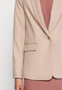 Blazer beige su misura con un singolo bottone dorato, rever a punta e tasca anteriore. Tessuto morbido, texture liscia, indossato sopra un vestito rosa chiaro.