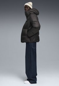 Sort pufferjakke med hætte, tykt quiltet design og logo på brystet; parret med løse mørke denimjeans og hvide sneakers.