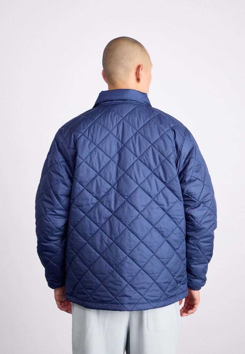 Veste matelassée marine avec un motif en diamant, coupe décontractée, col pointu et poignets élastiqués. Texture lisse, design minimaliste.