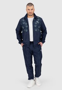 Marine tracksuit met een zipjack dat een geometrisch patroon heeft op het bovenste gedeelte, broek met elastische tailleband en witte sneakers.