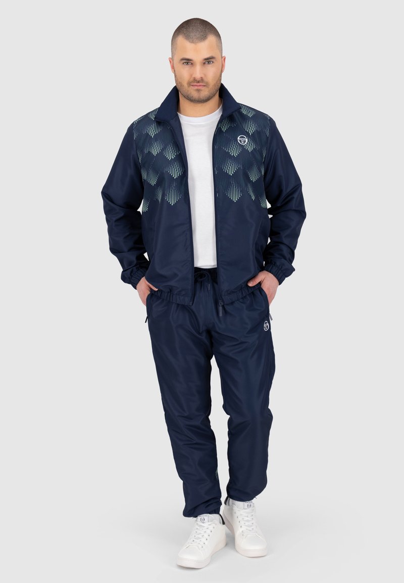 Marine tracksuit met een zipjack dat een geometrisch patroon heeft op het bovenste gedeelte, broek met elastische tailleband en witte sneakers.