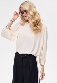 Jasnobeżowa bluzka z bufkami na rękawach, ściągaczami oraz miękką fakturą, w połączeniu z czarnymi spodniami z wysokim stanem i lampasami po bokach.