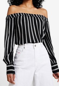 Blouse rayée noir et blanc sans épaules avec des manches longues, fabriquée en tissu léger, associée à un pantalon blanc taille haute.