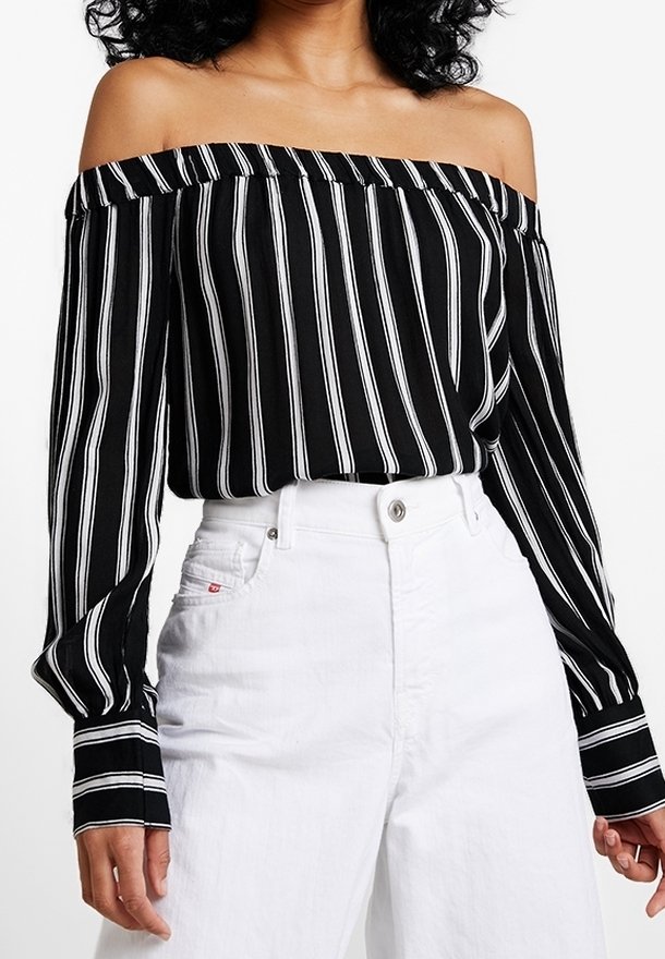 Blouse rayée noir et blanc sans épaules avec des manches longues, fabriquée en tissu léger, associée à un pantalon blanc taille haute.