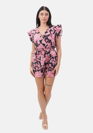 Vrouw met lang donker haar, gekleed in een zwarte en roze bloemenprint wikkelromper met ruches aan de mouwen en hakken met transparante bandjes, staand op een witte achtergrond.