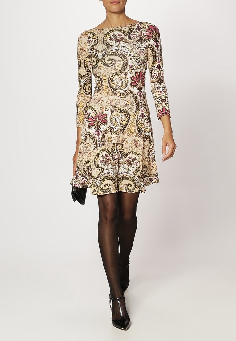Femme portant une robe longueur genou à manches longues avec un motif floral orné beige, noir et rouge, des collants noirs transparents et des talons noirs.